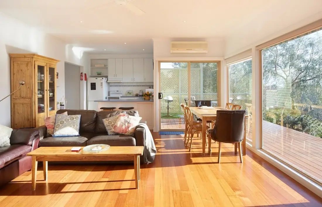 Open-plan living room with couches, dining table and kitchen, glass doors to the deck